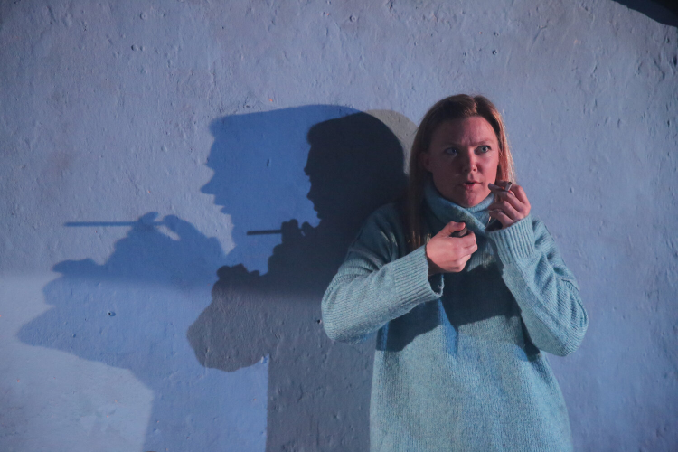 Actor in a blue jumper on stage in front of a white wall